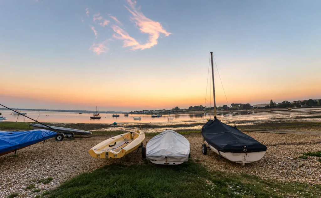 Professional sunset view of Mudeford Quay, showing small boats on the shingle shore, calm harbour water, moored boats in the distance and soft golden light across the sky.