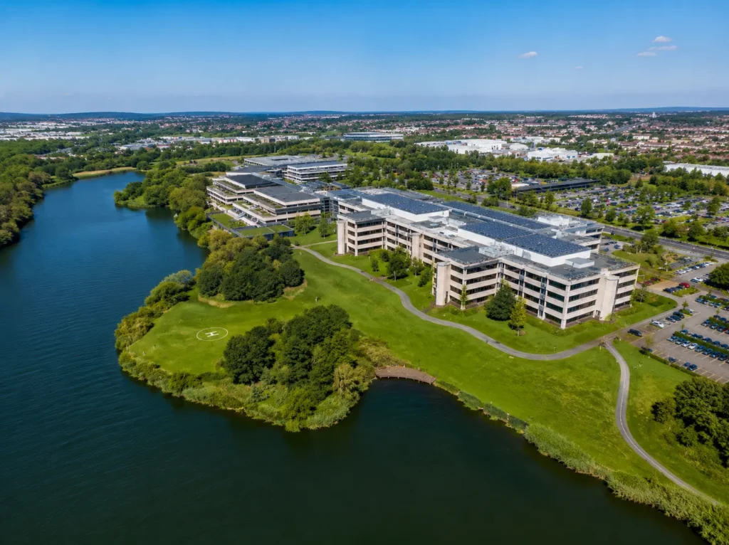 Aerial view of Lakeside North Harbour in Cosham, showing the modern office campus, solar-panelled rooftops, lakeside lawns, helipad, curved paths, car parks and surrounding water on a bright sunny day.