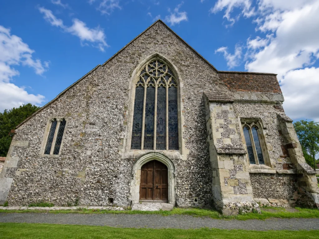 Professional exterior view of St Laurence’s Church, showing the flint-stone facade, large Gothic window, arched wooden doorway and bright blue sky with scattered clouds.
