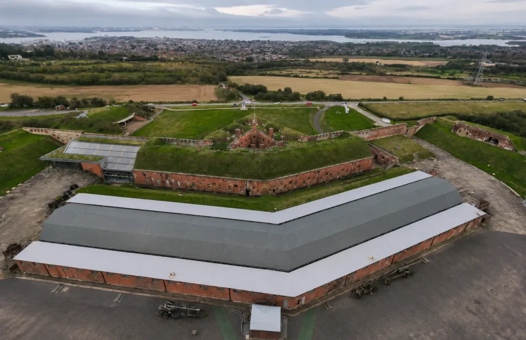 Aerial view of Fort Nelson in Fareham, showing the historic red-brick fort, grass-covered ramparts, museum buildings, surrounding fields and distant views across Portsmouth Harbour under an overcast sky.