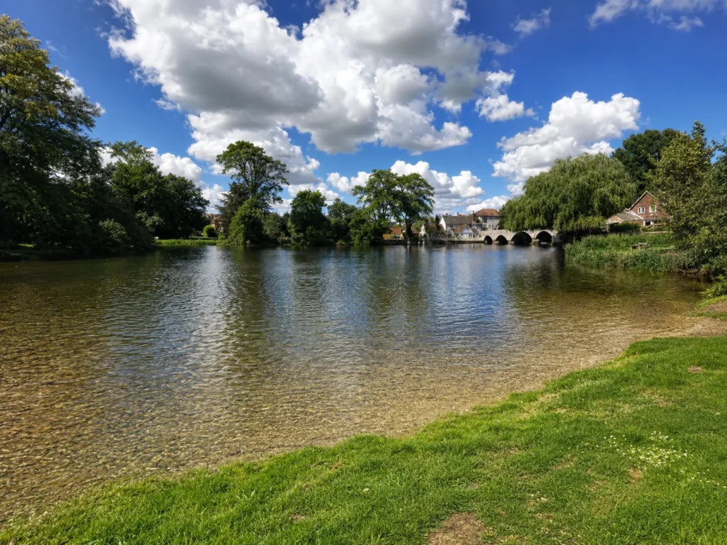 Riverside view in Ringwood, showing clear shallow water, grassy banks, mature trees, riverside homes and a stone bridge beneath a bright blue sky with clouds.