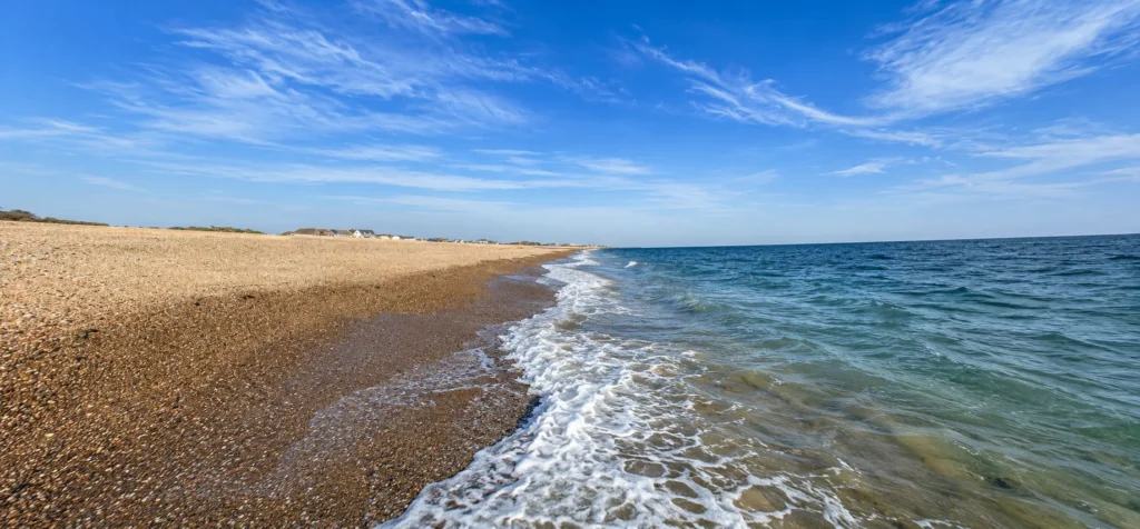 Professional wide-angle view of Sandy Point beach, showing the shingle shoreline, gentle waves, blue sea and a bright sky with wispy clouds.
