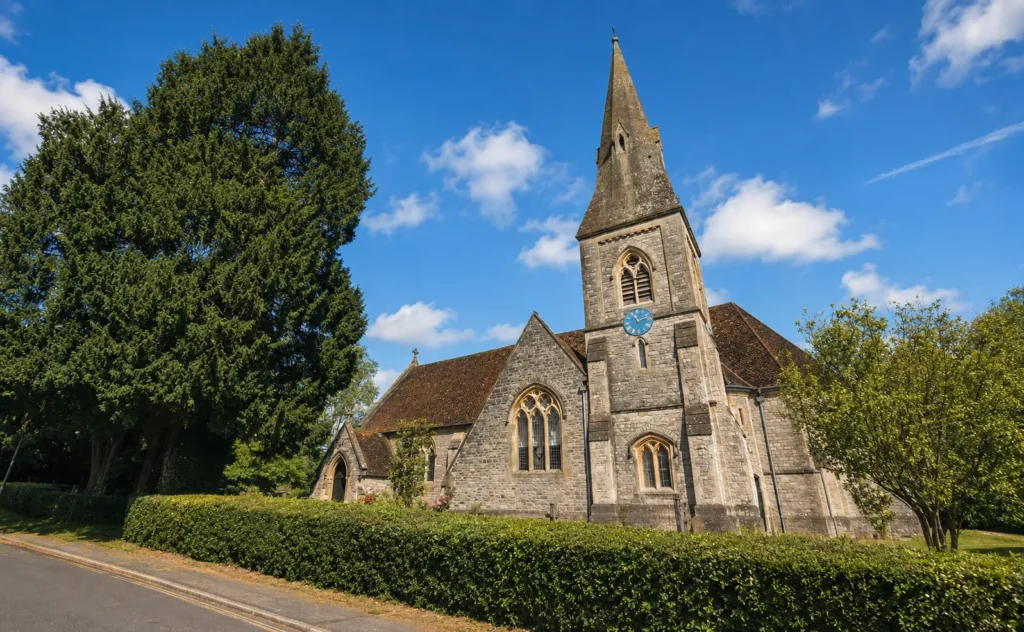 Sunny daytime view of St John’s Church in Hedge End, with its stone spire, blue clock face, mature trees, hedges and a few clouds in a bright blue sky.