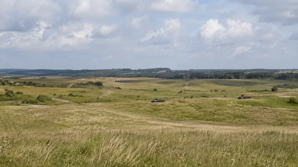 Wide Salisbury Plain-style landscape near Larkhill, showing open chalk grassland, MOD tracks, distant treelines and several military tanks moving across the fields under a cloudy sky.