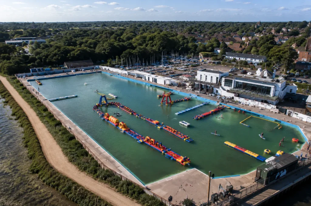 Aerial view of Lymington Sea Water Baths on a sunny day, showing the large open-air seawater pool, colourful inflatable obstacle courses, paddleboard areas, marina buildings and surrounding trees near the waterfront.