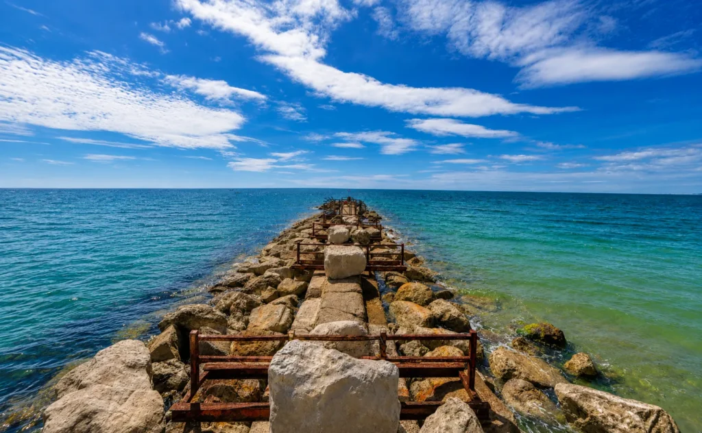 Professional view of Hengistbury Head Beach, showing a rocky groyne stretching into the sea, clear blue-green water and a bright sky with scattered clouds.