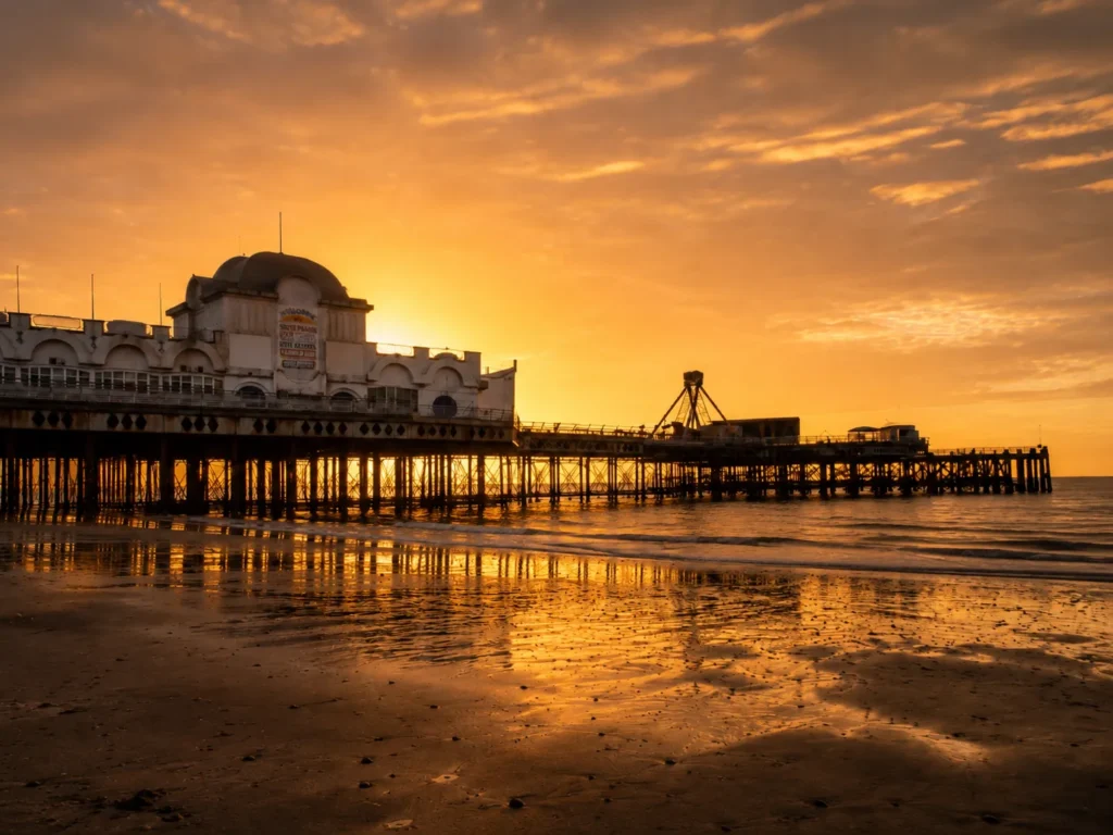 Sunrise view of South Parade Pier in Southsea, with the sun glowing behind the white pier building, golden light reflecting across the wet beach and waves beneath the pier.