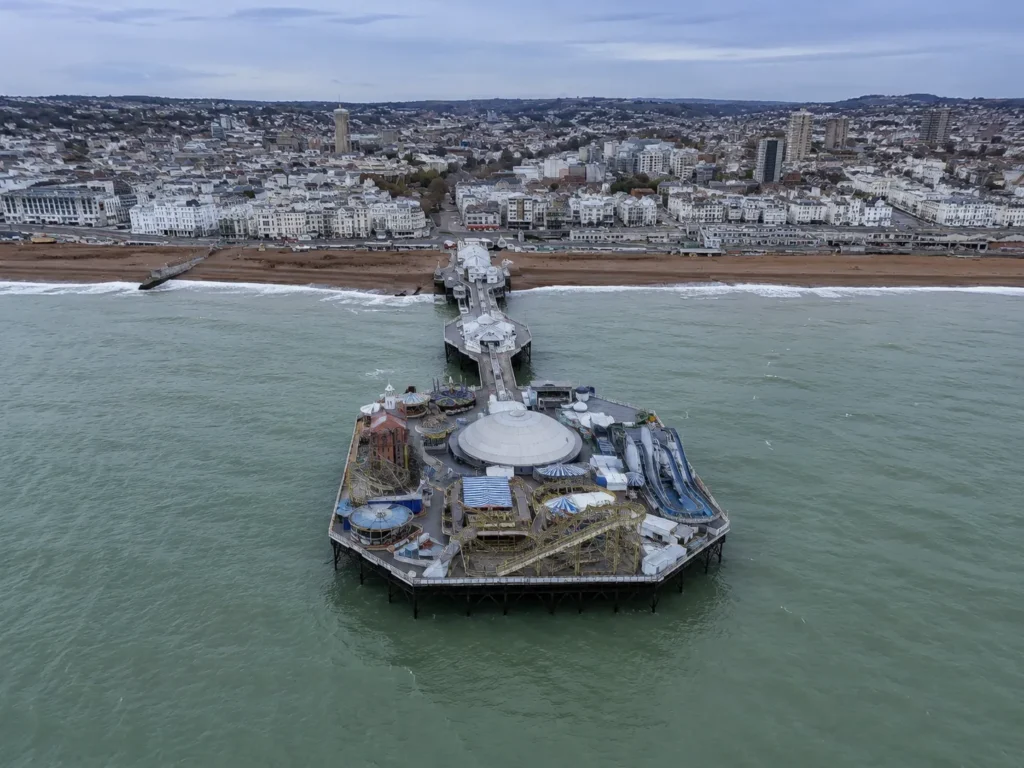Professional aerial view of Brighton Palace Pier and seafront, showing the pier’s domed building, beach, waves and the city skyline beneath an overcast sky.