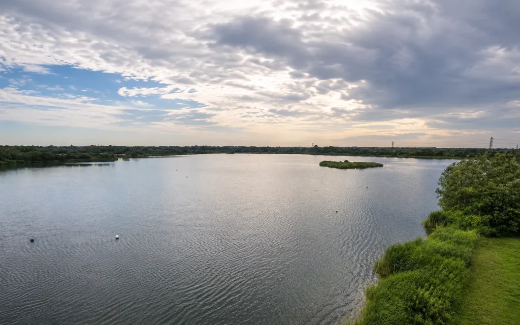 Professional wide-angle view of Longham Lakes in Ferndown, showing calm water, grassy banks, a small island, surrounding trees and a dramatic cloudy sky.