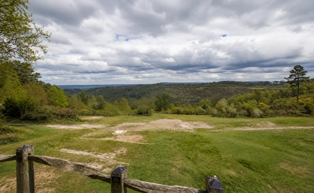 Panoramic view across the Devil’s Punch Bowl in Hindhead, with rolling heathland, wooded slopes and a dramatic cloudy sky seen from a fenced viewpoint.