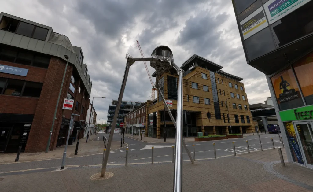 The Martian sculpture in Woking town centre beneath a dramatic overcast sky, surrounded by modern buildings and quiet streets.